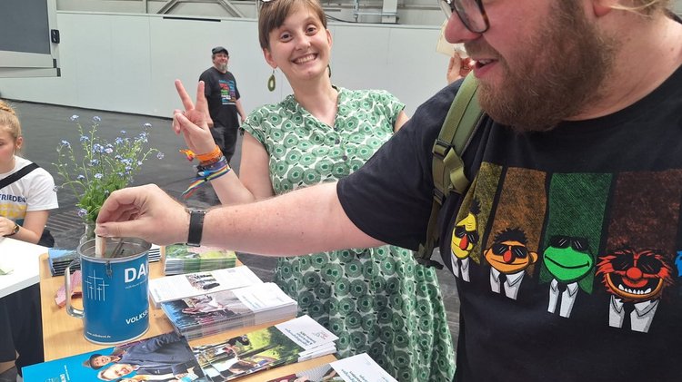 Ein Mann steckt einen Geldschein in die Spendendose des Volksbundes. Die Frau im Hintergrund zeigt das Victory-Zeichen.