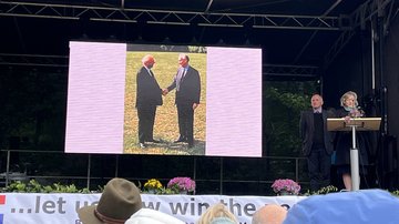 Eine Frau und ein Mann auf der Bühne. Im Hintergrund ein Foto von zwei Männern die sich die Hand geben.