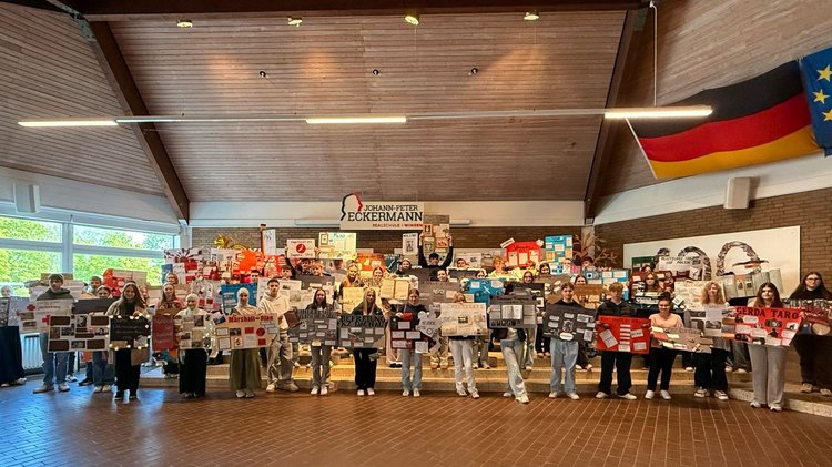 Eine große Gruppe von Schülerinnen und Schülern in einer Halledie Plakate in den Händen halten. Über der Gruppe das Logo der Schule. 