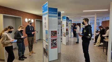 In einem breiten Gang sind Ausstellungswände der Ausstellung „Zeitenwende ’45 – Aufbruch in ein neues Europa“ hintereinander aufgestellt. Links und rechts davon Schülerinnen und Schüler. Rechts auf einen Tisch sitzen weitere Jugendliche. Alle haben Tablets in den Händen und tragen Masken.