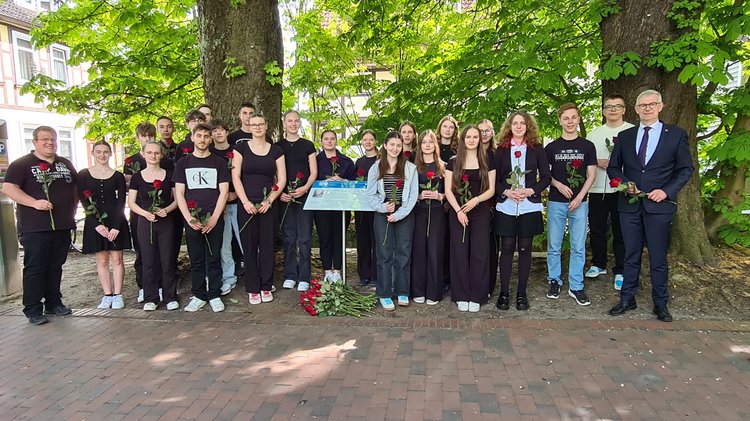Jugendliche und zwei erwachsene Männer stehen um die Geschichts- und Erinnerungstafel. Alle halten eine rote Rose in den Händen. Vor der tafel liegen weitere Rosen.