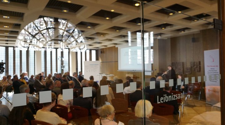 Blick durch eine Glastür des Leibnizhauses in einen Saal in dem viele Menschen sitzen