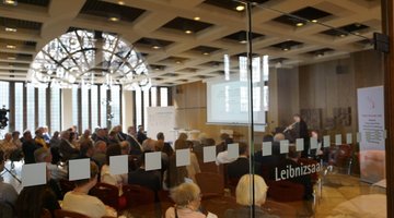 Blick durch eine Glastür des Leibnizhauses in einen Saal in dem viele Menschen sitzen