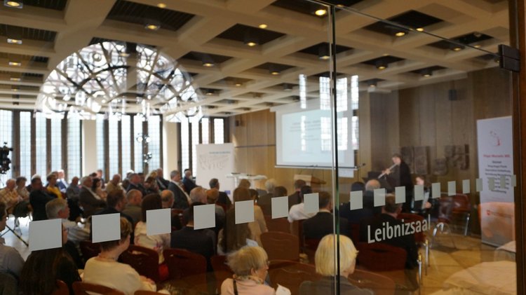 Blick durch eine Glastür des Leibnizhauses in einen Saal in dem viele Menschen sitzen