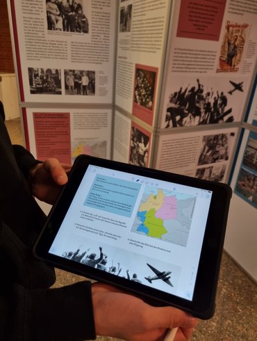 Im Vordergrund halten Hände ein Tablet in die Kamera. Darauf zu sehen, ein Ausschnitt aus einem Fragebogen für Schülerinnen und Schüler zur Ausstellung „Niedersachsen im Krieg“. Ausstellungswände hiervon sind im Hintergrund zu sehen.