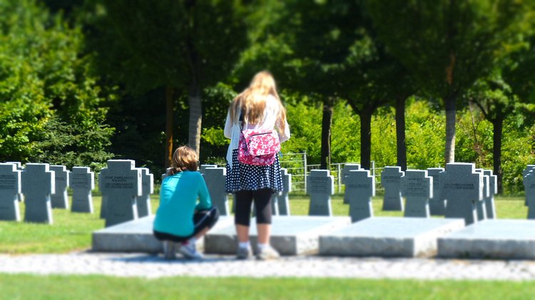 Zwei weibliche Jugendliche auf einer Kriegsgräberstätte vor einer Grabplatte die sie betrachten. Im Hintergrund Grabsteine.