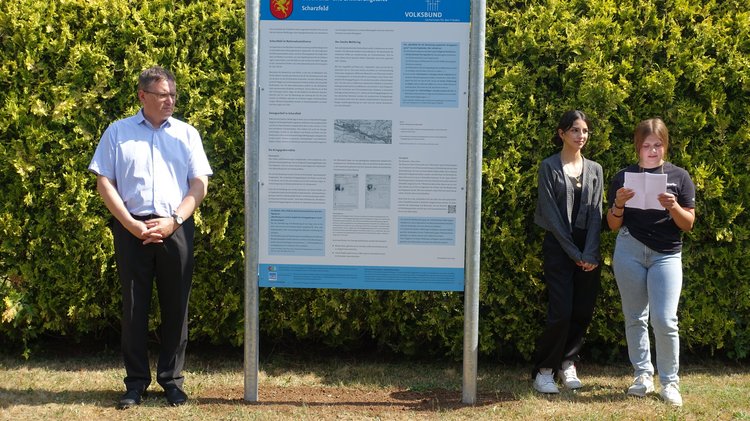 Zwei Schülerinnen stehen neben der Geschichts- und Erinnerungstafel auf dem Friedhof Scharzfeld. Eine trägt einen Text von einem Blatt Papier vor.