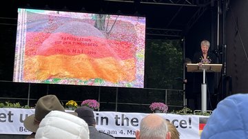 Eine Frau steht hinter dem Rednerpult auf der Bühne und spricht in das Microfon. Auf der Leinwand die Deutsche Flagge.