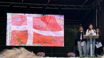 Drei Schülerinnen auf der Bühne. Ein Mädchen spricht in das Microfon. Auf einer Leinwand die Dänische Flagge.