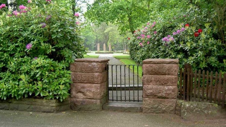 Eingangstor zur Kriegsgräberstätte Sandbostel. Links und rechts blühende Büsche, im Hintergrund Gedenkstelen.