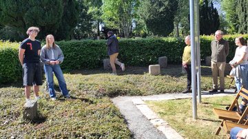 Links eine Schülerin und ein Schüler. Die Jugendliche spricht. Rechts, hiner der Tafel, Zuhörende.