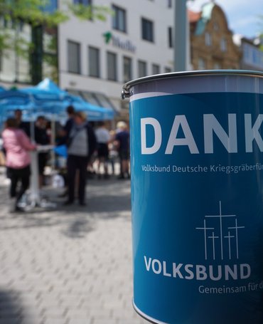Vorne rechts, leicht angeschnitten, eine Spendendose des Volksbundes. Im Hintergrund, verschwommen, Menschen an einem Infostand des Volksbundes