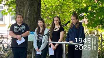 Drei Schülerinnen und ihr Lehrer. Eine der Jugendlichen spricht in ein Mikrofon.