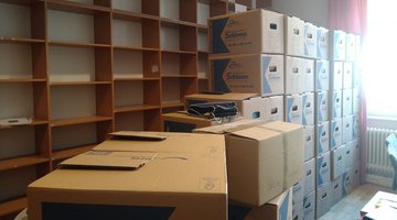 Ein großer Stapel Umzugskartons. Dahinter ein langes, hohes Regal das leer ist.