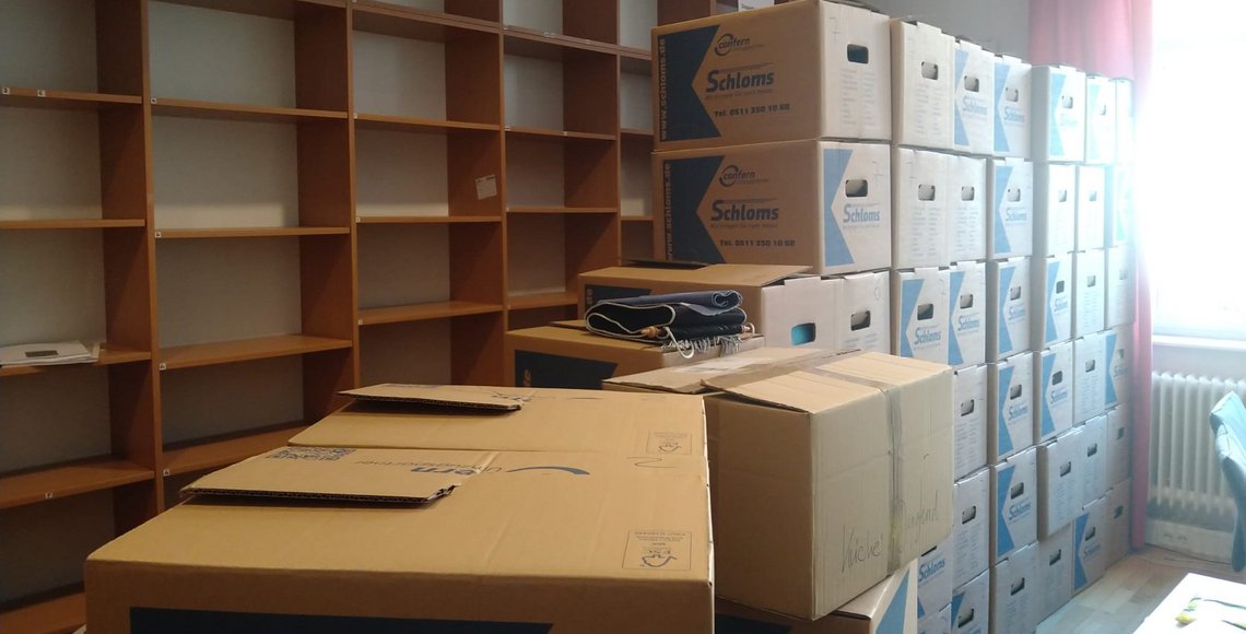 Ein großer Stapel Umzugskartons. Dahinter ein langes, hohes Regal das leer ist.