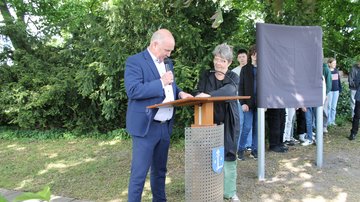 Ein Mann spricht in eine Mikrofon, daneben eine Frau. Im Hintergrund die verhüllte Geschichts- und Erinnerungstafel, dahinter Schülerinnen und Schüler.