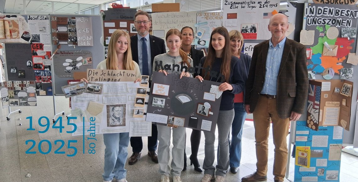 Eine Gruppe von Schülerinnen stehen zusammen mit zwei Männern und einer Frau vor Ausstelungswänden. Sie halten selbstgestaltete Ausstellungsplakate in den Händen.