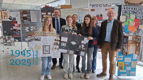 Eine Gruppe von Schülerinnen stehen zusammen mit zwei Männern und einer Frau vor Ausstelungswänden. Sie halten selbstgestaltete Ausstellungsplakate in den Händen.